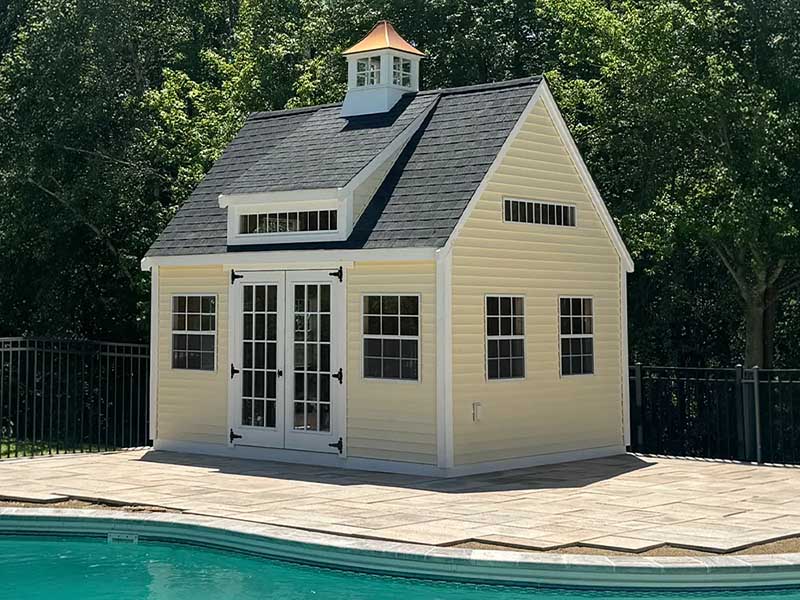 yellow backyard shed next to a pool with a dark gable roof, lots of windows and double French doors