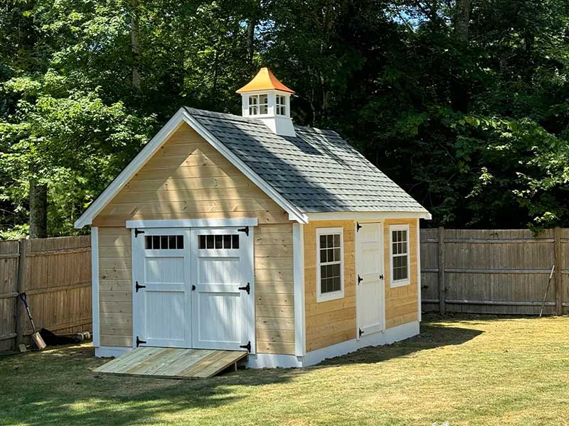 yellow backyard shed with a black pitch roof and decorative elements in a grassy backyard with a fence