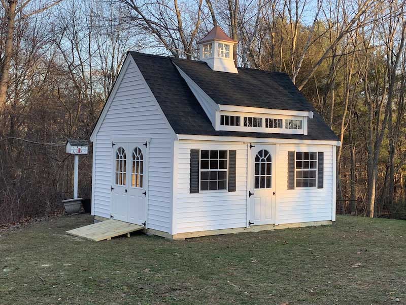 white backyard shed with lots of adornments, installed in a backyard with trees and other outdoor elements